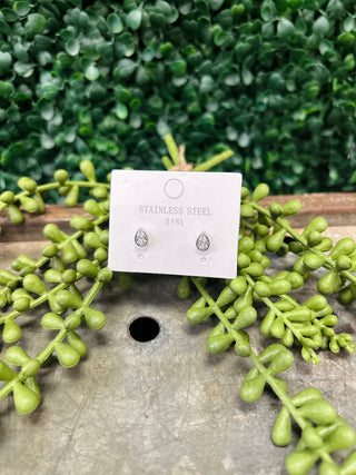 Pear-cut stud earrings on white packaging card surrounded by green foliage on wooden surface