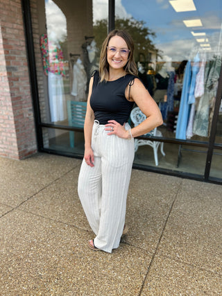 Woman wearing black sleeveless top and white pinstripe linen pants standing outside clothing boutique storefront.