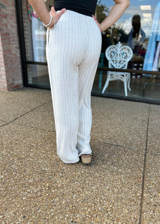 Woman wearing white pinstripe linen pants with pockets, standing on concrete sidewalk outside storefront.
