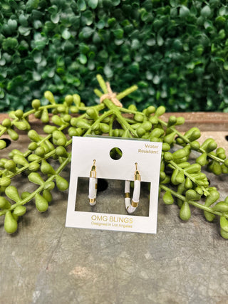 White and gold hoop earrings on display card surrounded by green foliage on wooden surface.