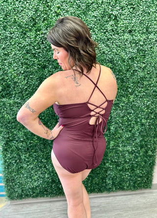 Woman wearing plum one-piece swimsuit with cross-back lacing detail, standing against green backdrop.