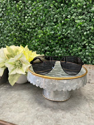 Black polarized aviator sunglasses displayed on decorative glass pedestal with flowers and greenery backdrop.