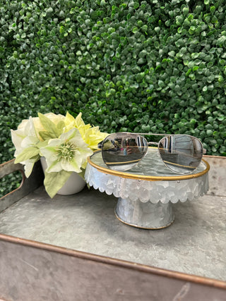 Polarized sunglasses with gold frames resting on decorative cake stand next to white flowers on marble surface.