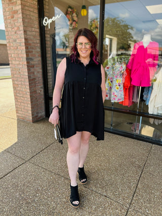 Woman wearing black sleeveless button-up dress with gathered waist, standing outside clothing boutique storefront.