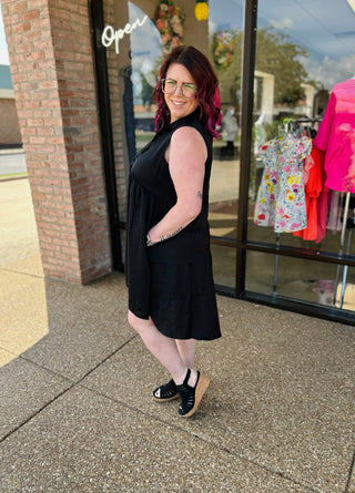 Woman wearing black sleeveless dress standing outside clothing boutique storefront with brick columns and glass windows.