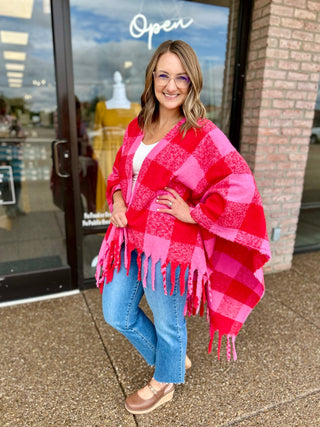 Woman wearing a fringed plaid wrap poncho, styled with jeans and clogs, posing outside a boutique.