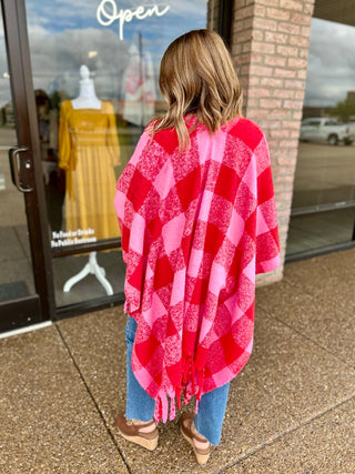 Woman wearing a plaid fringe wrap poncho, shown from behind outside a boutique storefront.
