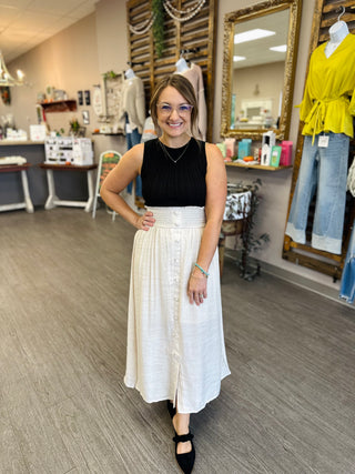 Woman wearing a sleeveless ribbed knit sweater tank paired with a white midi skirt in a boutique.