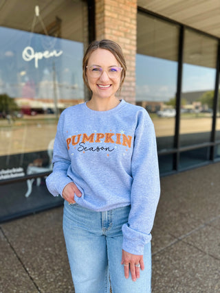 Woman smiling outdoors wearing a heather grey "Pumpkin Season" embroidered crewneck sweatshirt with jeans.