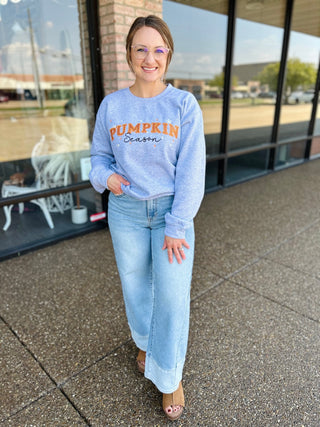 Woman wearing a heathered crewneck sweatshirt with embroidered "Pumpkin Season" graphic lettering outdoors.