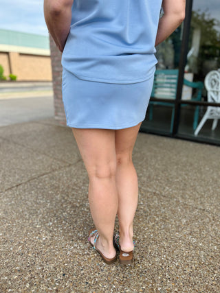 Woman wearing light blue athletic skort and sandals standing on pavement, rear view showing fit and style.