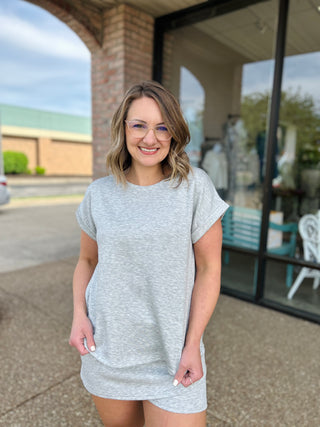 Woman wearing grey short-sleeve casual t-shirt dress standing outdoors in front of brick building
