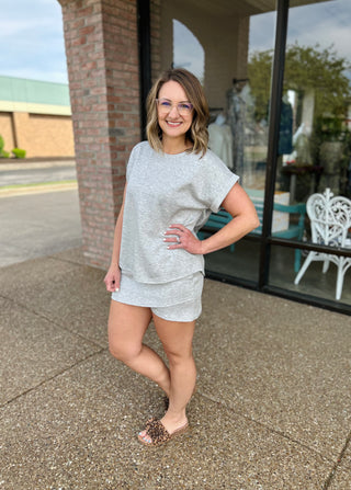 Woman wearing grey casual short-sleeve top and matching shorts, standing outside storefront with glasses and sandals.