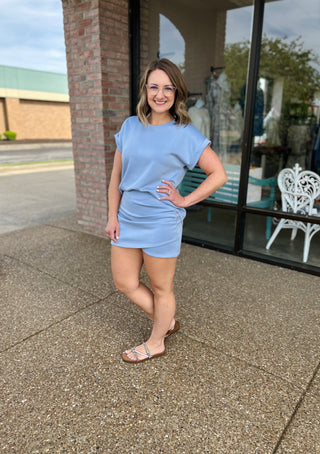 Woman wearing light blue short-sleeve top and matching skirt, standing outdoors near storefront