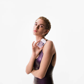 Model holding Touchland Pure Lavender Power Mist hand sanitizer against her shoulder, wearing purple bodysuit.