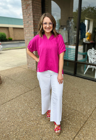 Woman wearing bright pink collared blouse with white pants and colorful sandals, standing outdoors smiling.