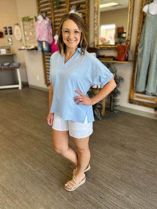 Woman wearing light blue collared blouse with white shorts and woven sandals in clothing boutique