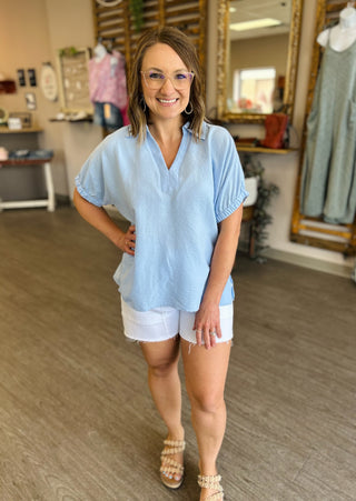 Woman wearing light blue chambray collared blouse with white shorts in retail store setting.