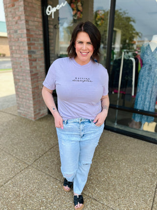 Woman wearing purple "Raising Disciples" embroidered t-shirt with jeans standing outside boutique storefront