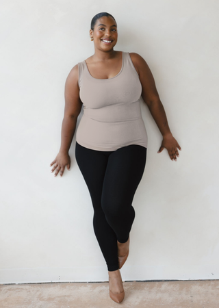 Woman wearing beige reversible cami tank top with black leggings, smiling against white wall.