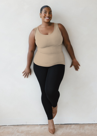 Woman wearing beige reversible cami tank top with black leggings, smiling against white wall.