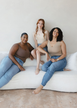 Three women wearing neutral-toned reversible camisoles and jeans sitting casually on white cushions