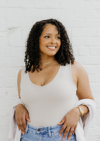 Woman wearing beige reversible cami top with white cardigan and jeans against brick wall background.