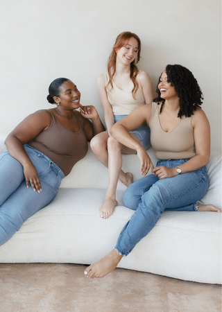 Three diverse women wearing neutral-toned reversible camisoles and jeans sitting together on white cushions, smiling.