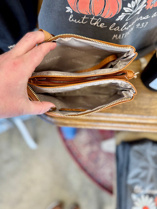 Hand holding open tan leather crossbody wristlet showing three zippered compartments with gray fabric lining