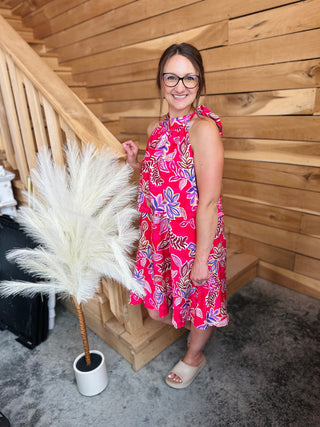 Woman wearing floral sleeveless mini dress with tie shoulders, standing by wooden staircase with pampas grass decor.