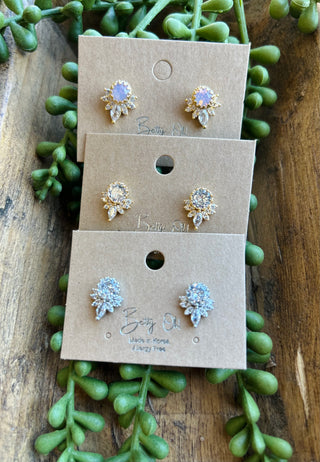 Three pairs of sparkly rosette stud earrings displayed on kraft paper cards with green succulent backdrop.