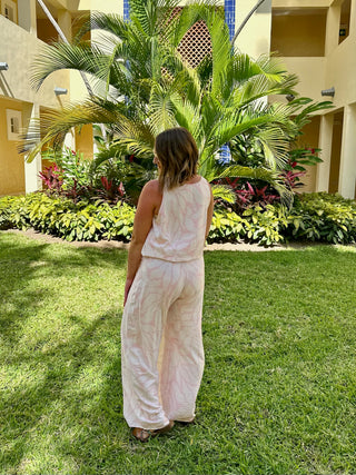Woman wearing flowing palazzo pants and sleeveless top standing on grass near tropical landscaping.