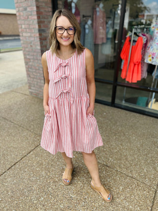 Woman wearing pink and white striped sleeveless dress with tie details standing outside boutique storefront.
