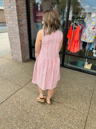 Woman wearing pink and white striped sleeveless dress with tie detail, standing outside boutique storefront.