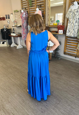 Woman wearing blue sleeveless tiered maxi dress with elastic waist, shown from behind in clothing boutique.
