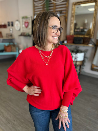 Woman wearing red relaxed dolman sweater with layered gold necklaces, smiling in casual indoor setting.