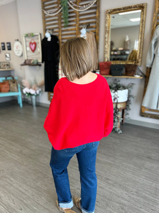 Woman wearing red oversized dolman sweater with jeans in boutique setting, shown from behind