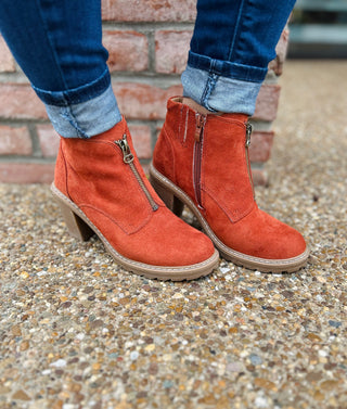 Person wearing rust-colored faux suede ankle boots with zippers and rolled cuffed jeans on textured ground.