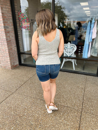 Woman wearing sage green knit tank top and denim shorts walking on sidewalk outside boutique storefront.