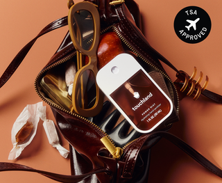 Touchland Power Mist hand sanitizer resting on an open brown leather handbag with sunglasses and caramel candies.