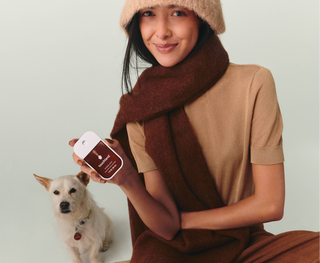 Woman in cozy autumn outfit holding Touchland Power Mist Hand Sanitizer beside a small white dog.