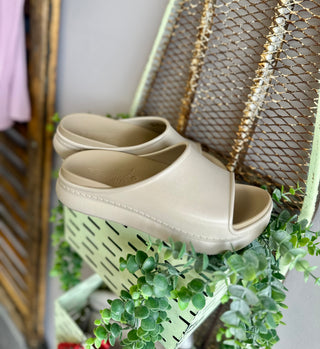 Dansko sand platform slide sandals displayed on mint green crate with decorative plants and woven basket.