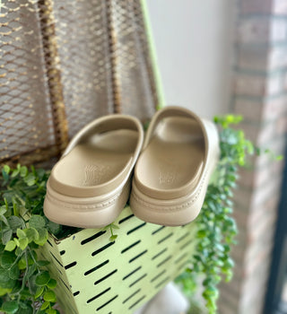 Beige platform slide sandals displayed on green metal basket with plants in outdoor setting.