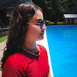 Woman wearing sunglasses and red top by swimming pool, showcasing delicate chain necklace from Alegria Accessories.