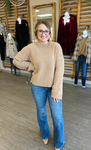 Woman wearing beige textured knit sweater with jeans in clothing boutique store setting.