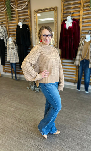 Woman wearing beige textured knit sweater with jeans, posing in clothing boutique with displayed outfits.