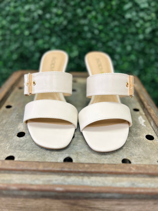 Sarika off white dress sandals with dual straps and gold buckles on wooden surface