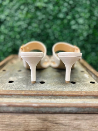 Pair of white dress sandals with heels displayed on wooden surface with green foliage background