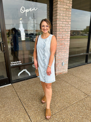 Woman wearing a sleeveless denim shift dress with decorative scalloped button-front detail, styled with heels.