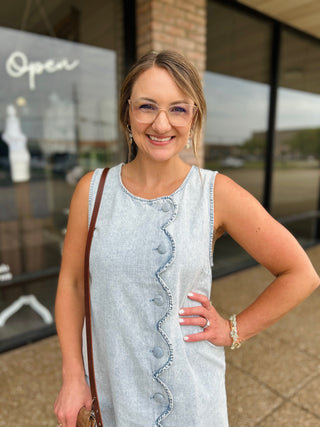 Woman wearing a sleeveless denim dress with scalloped button-front detail, styled with accessories.